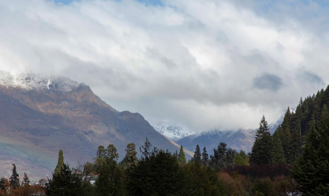 A moody mountain scene with clouds, trees, and snow.